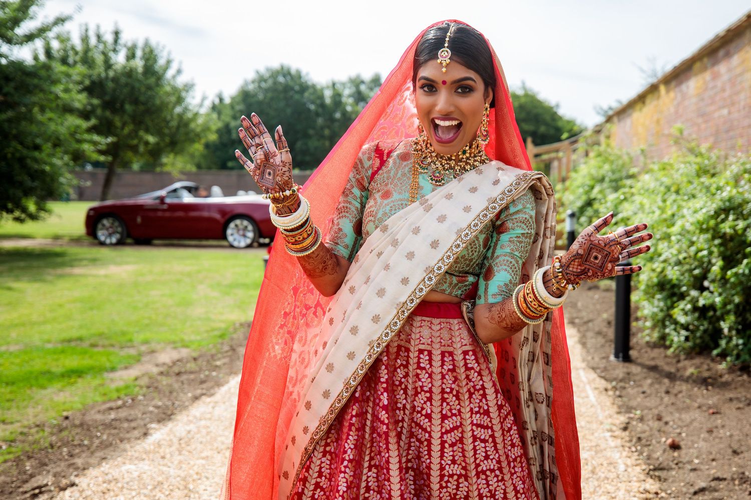 Traditional Indian bride in red lehenga poses outdoors with luxury car in background.