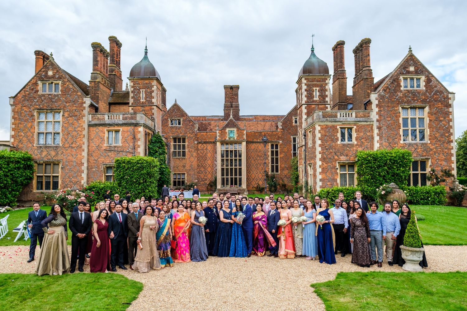 Large wedding party group photo in front of grand Tudor-style mansion with distinctive architecture.
