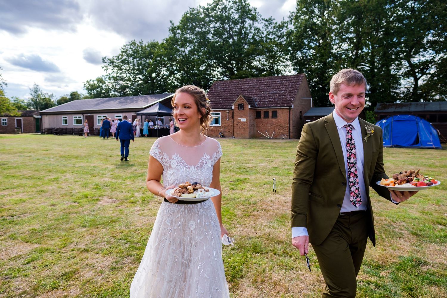 Wedding celebration taking place on a green field with brick buildings in the background.