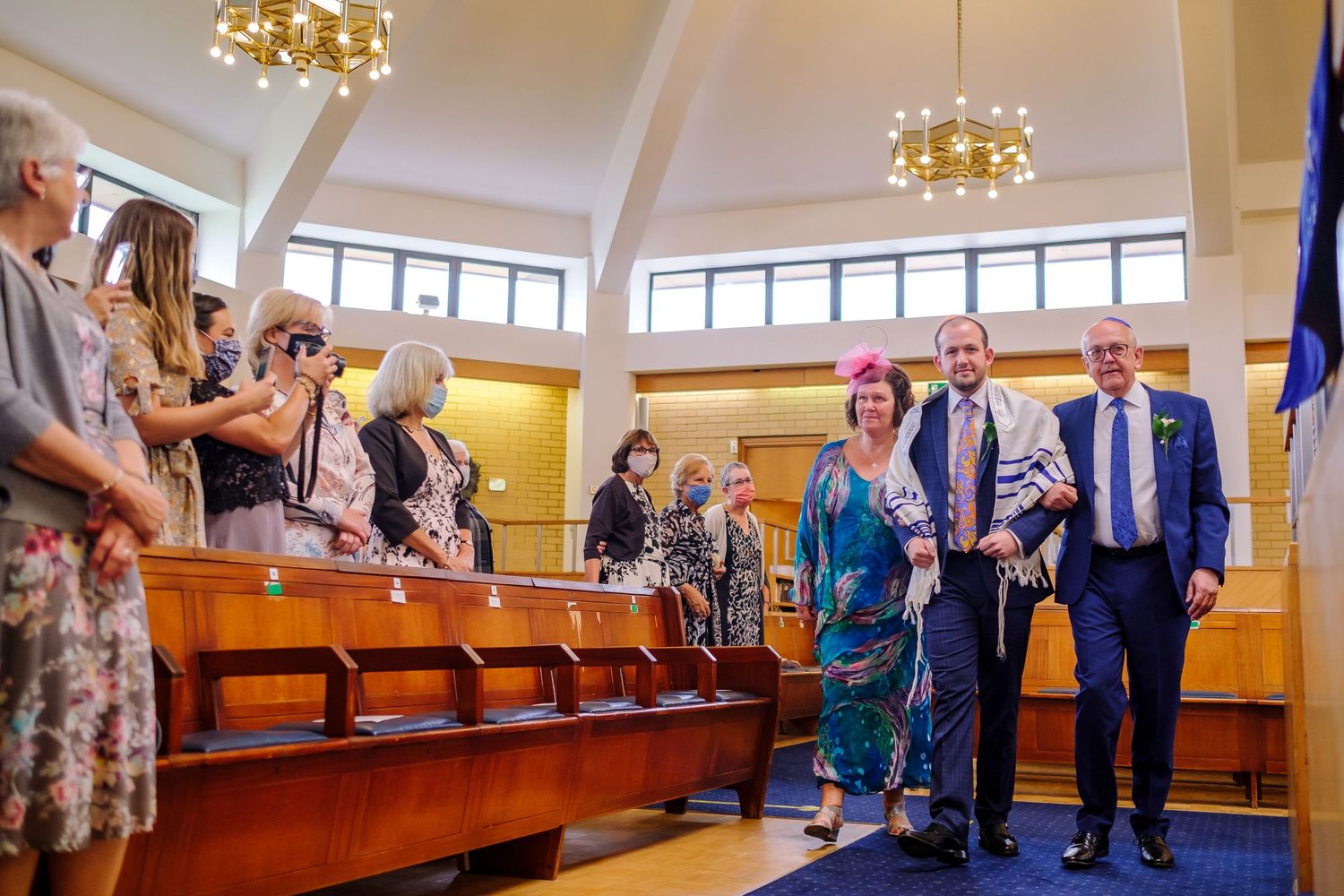 A group gathers in a bright chapel with wooden pews and golden chandeliers during a special ceremony.