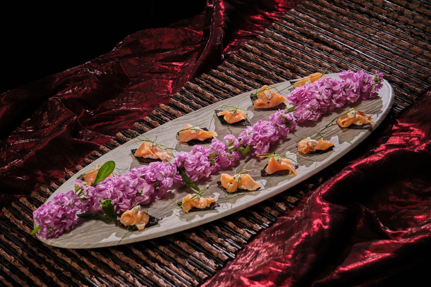 Elegant oval platter with purple flowers and smoked salmon bites on a woven surface with red fabric backdrop.