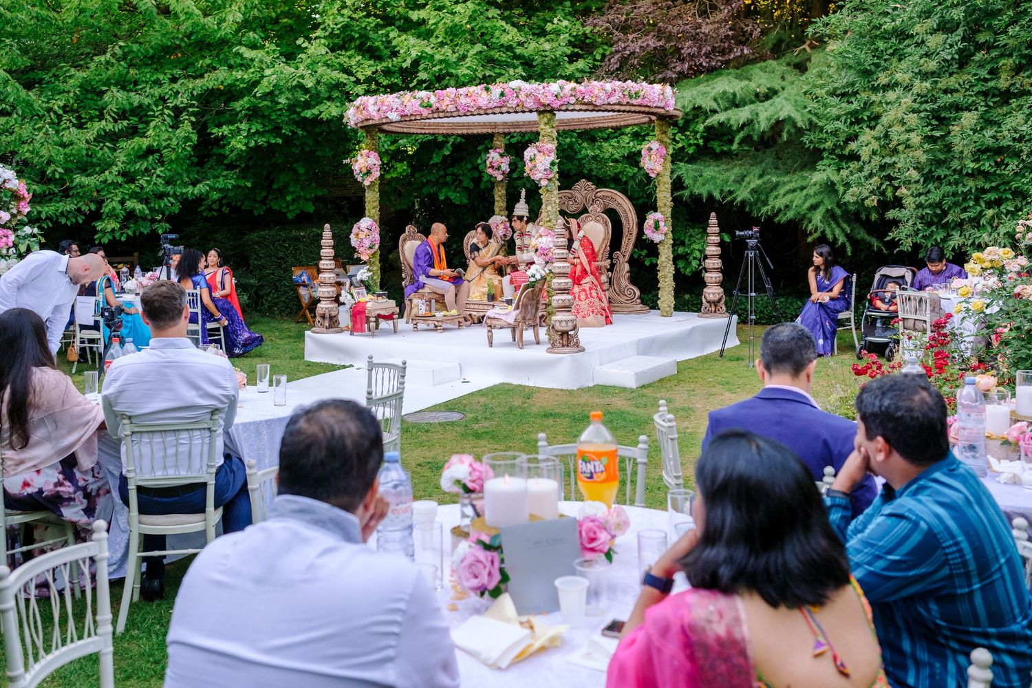 An outdoor wedding ceremony under a decorated wooden pergola surrounded by guests and greenery.