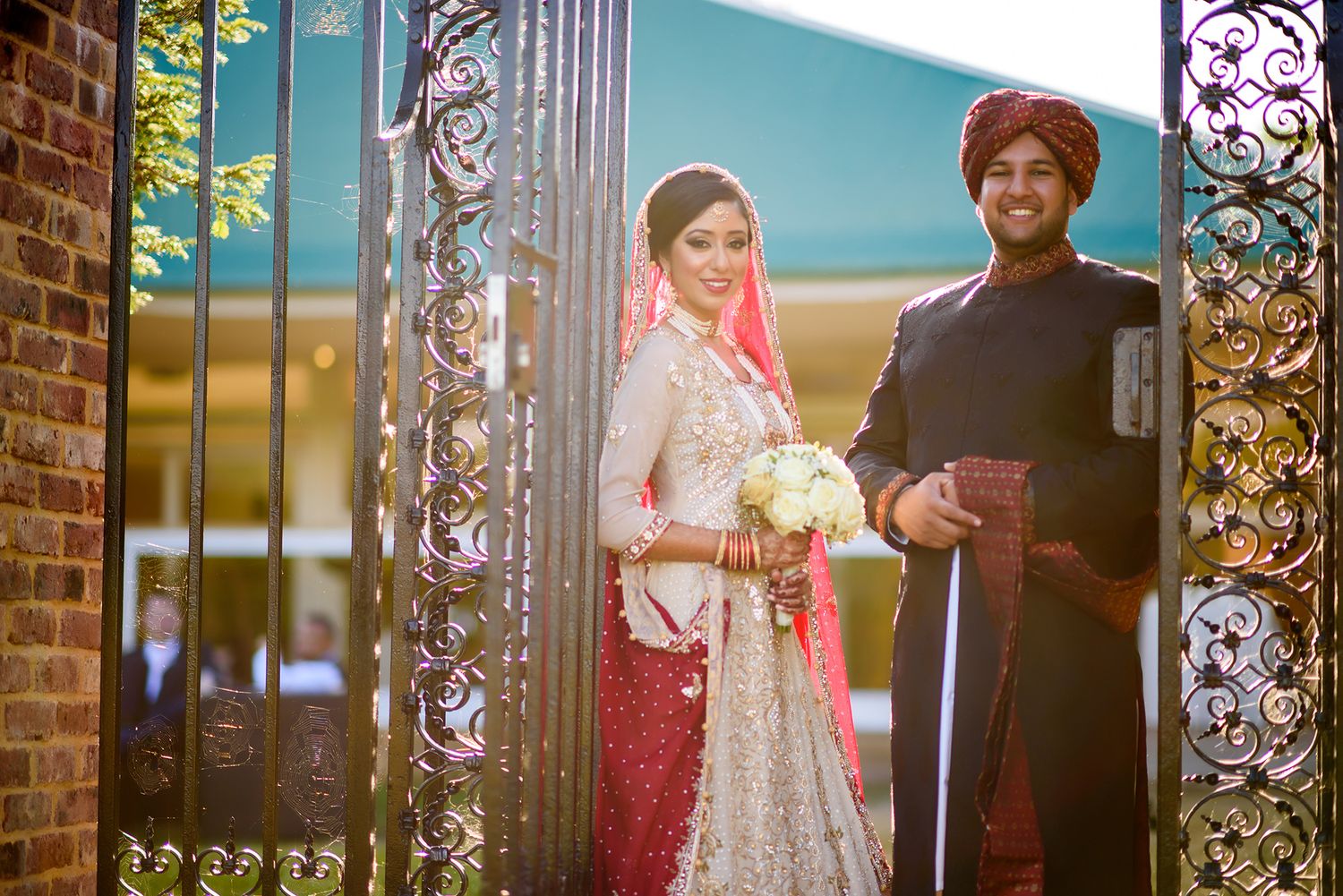 Traditional South Asian couple posing in ornate wedding attire by decorative gate at sunset.