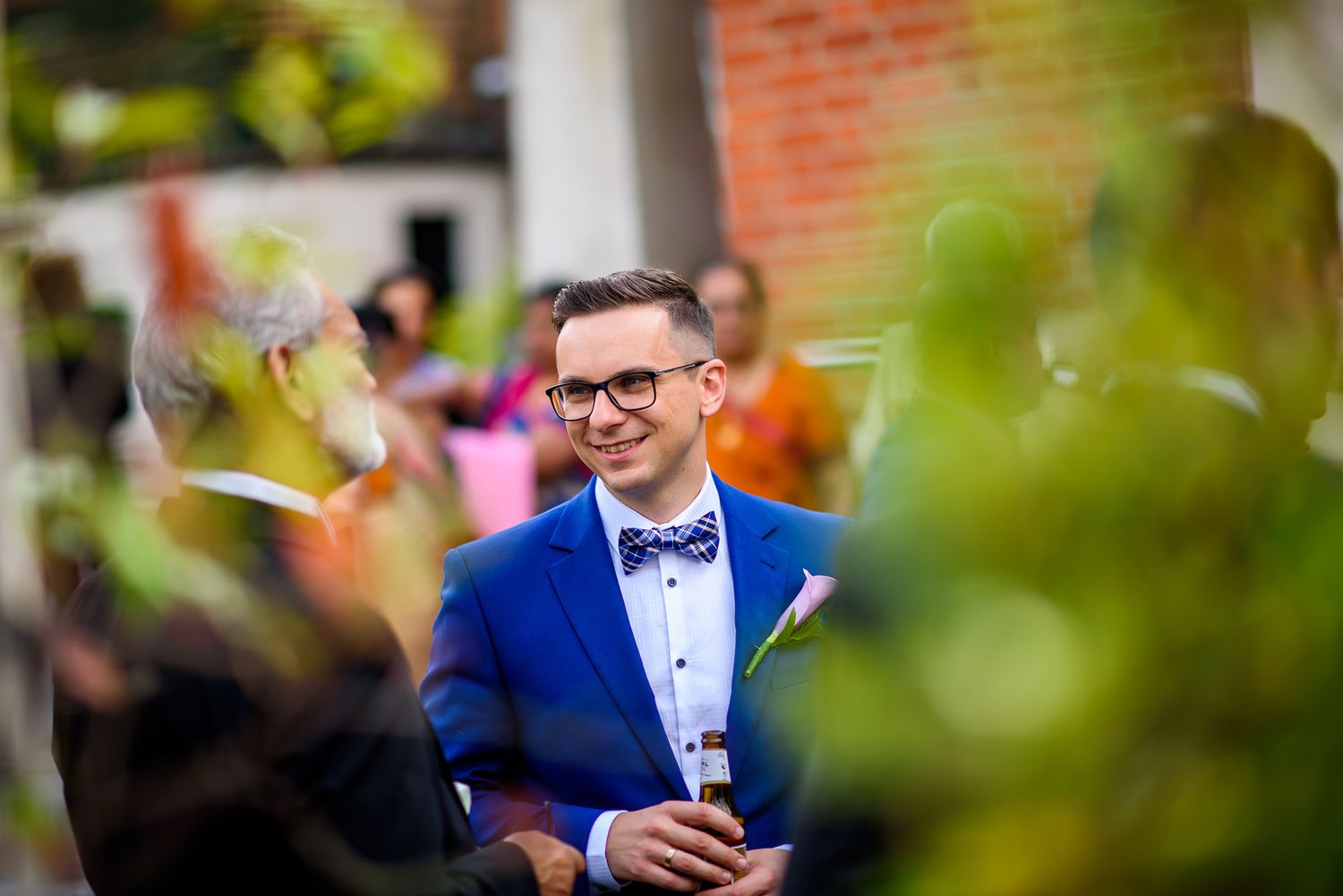 Guest in bright blue suit and bowtie enjoying the wedding celebration through garden foliage.