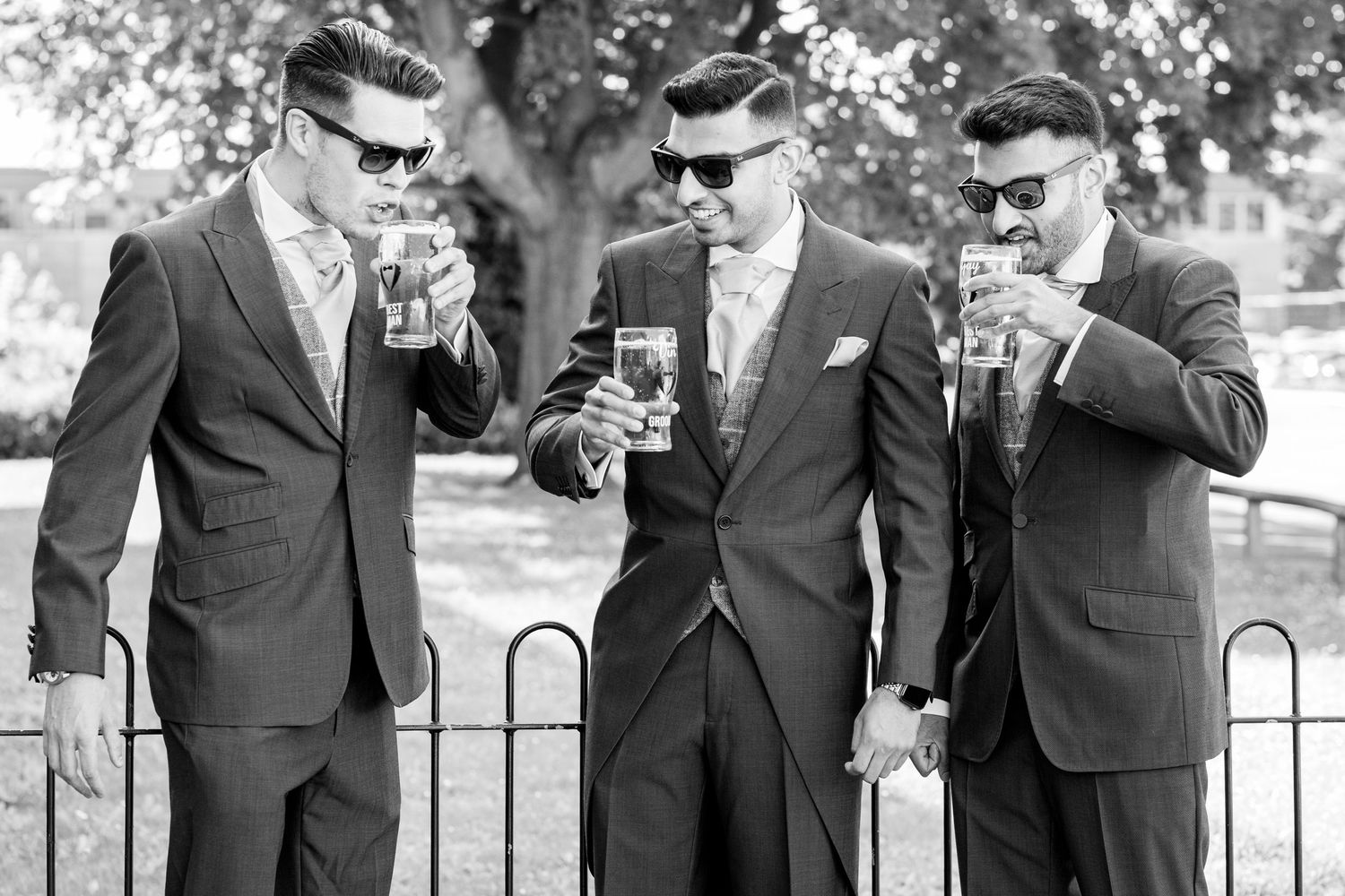 Groomsmen in dark suits stand together outdoors wearing sunglasses and holding drinks.