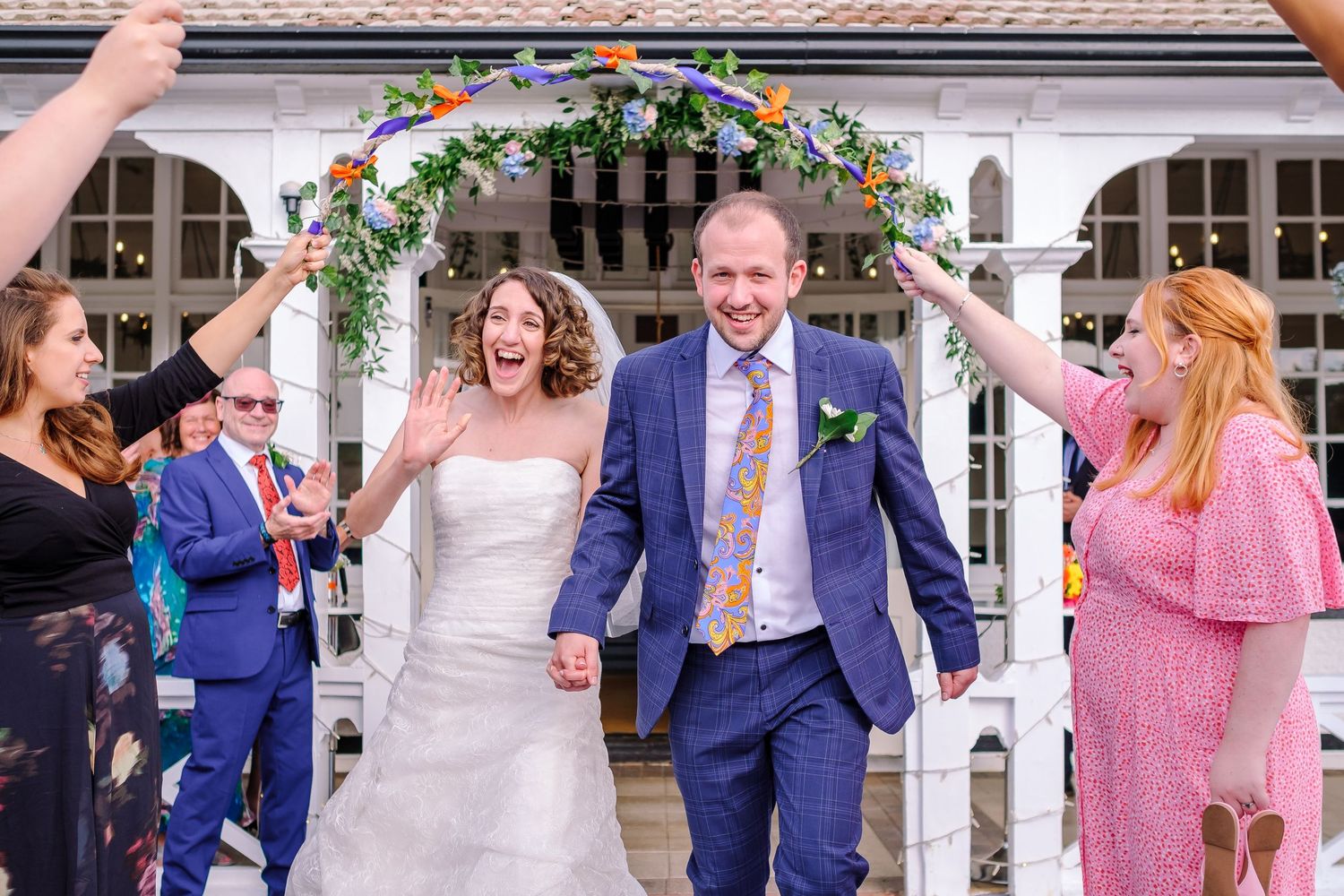 A couple exits their wedding ceremony under a floral arch while guests celebrate with confetti and bubbles.