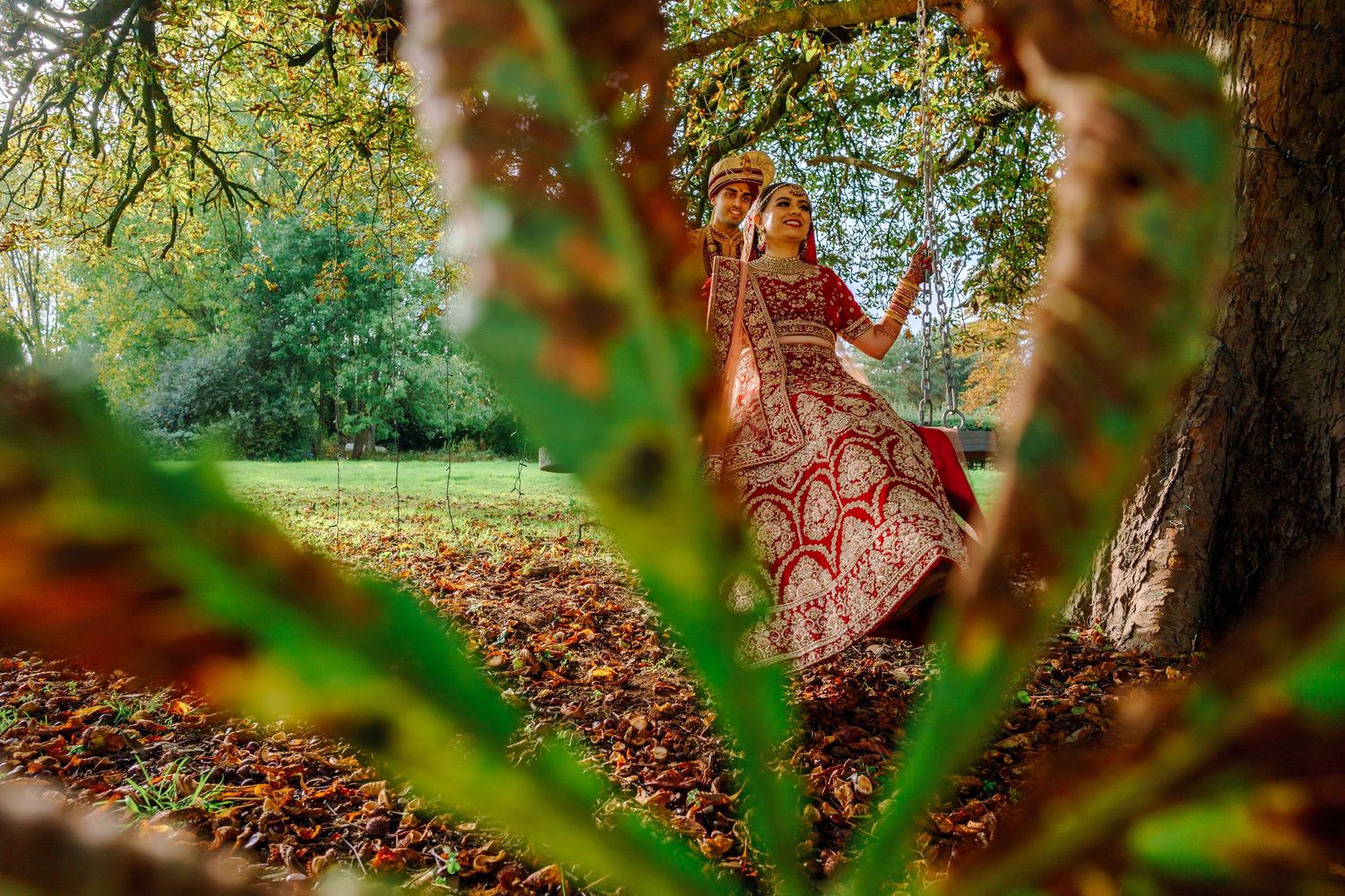 Artistic wedding photography through foliage showing traditional Indian bridal outfit in autumn setting.