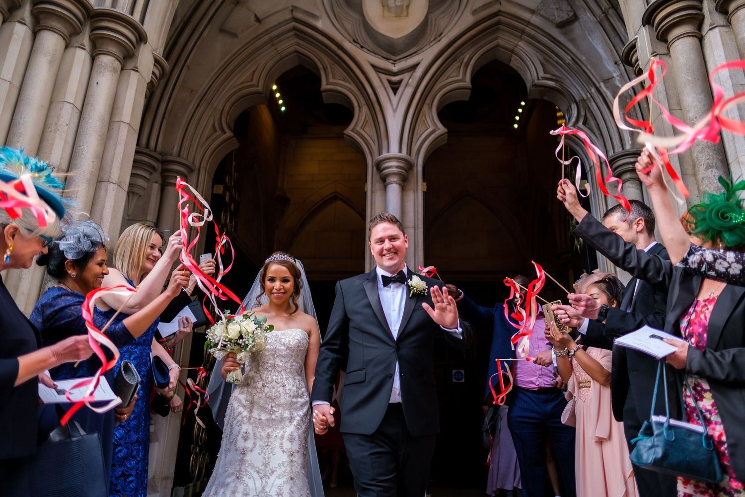 Wedding guests wave ribbons as newlyweds exit through stone archway after ceremony.