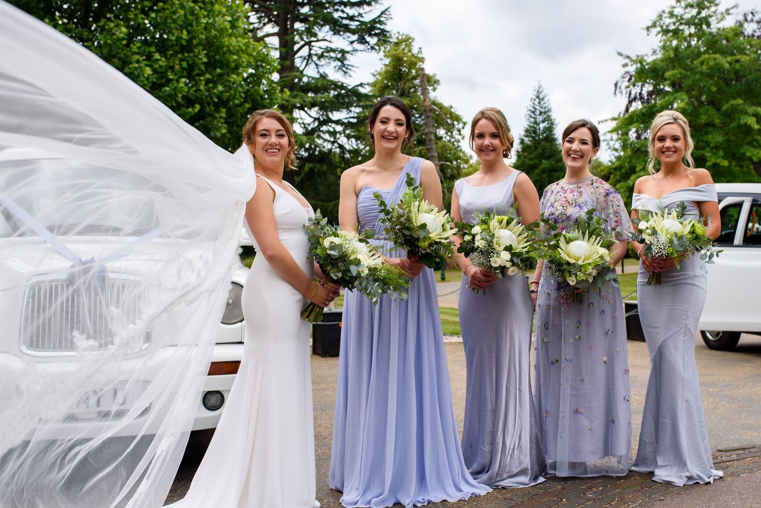 Wedding party in light purple bridesmaid dresses poses outdoors with flowing white veil in the breeze.