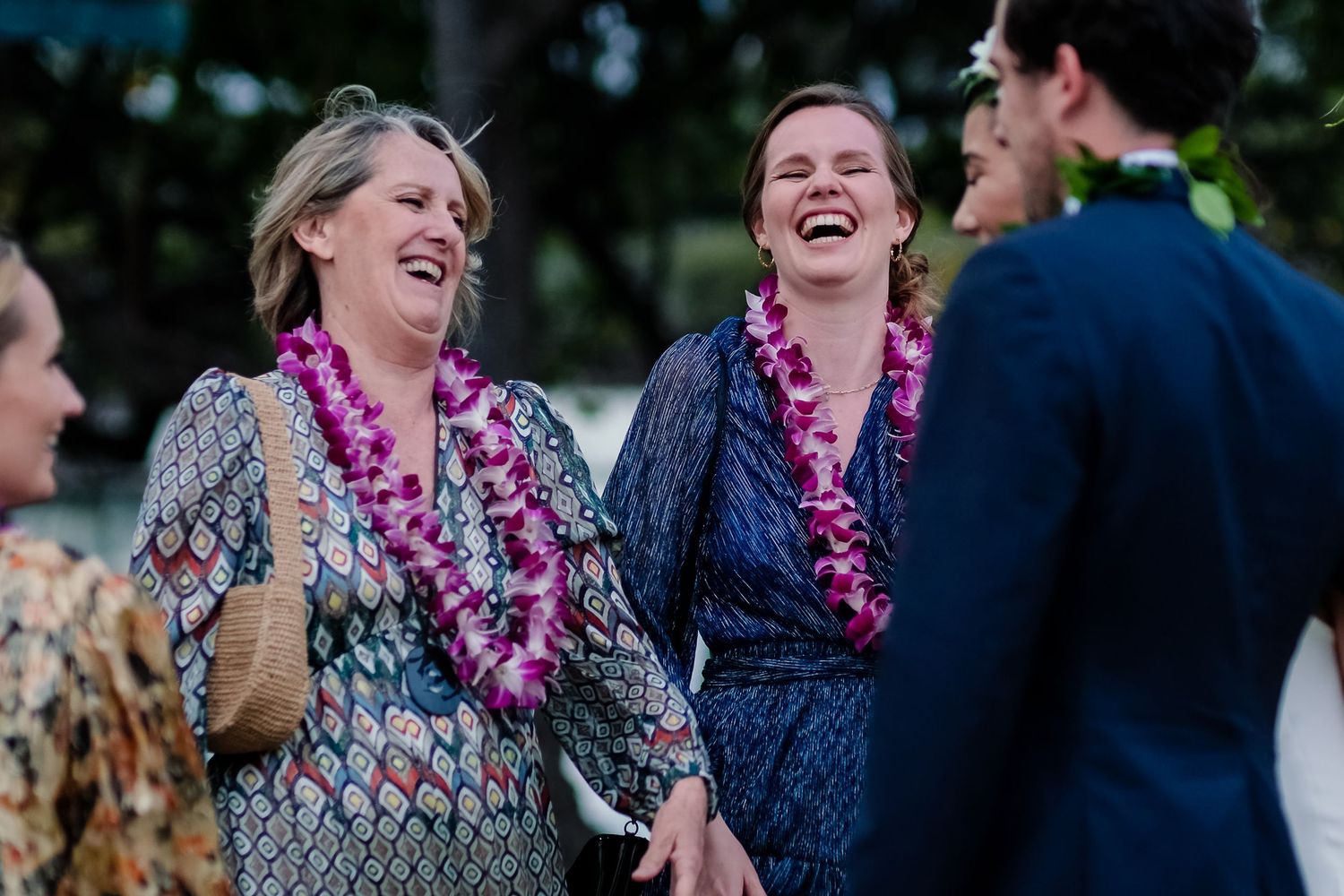 Wedding guests wearing purple leis laugh and smile during an outdoor celebration.