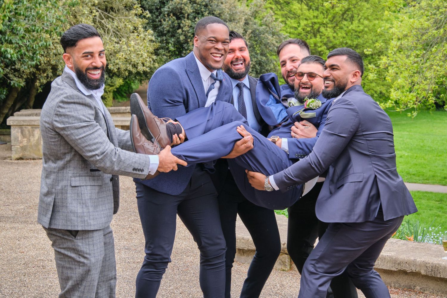 A playful group photo of groomsmen in suits lifting someone up while laughing together in a garden setting.