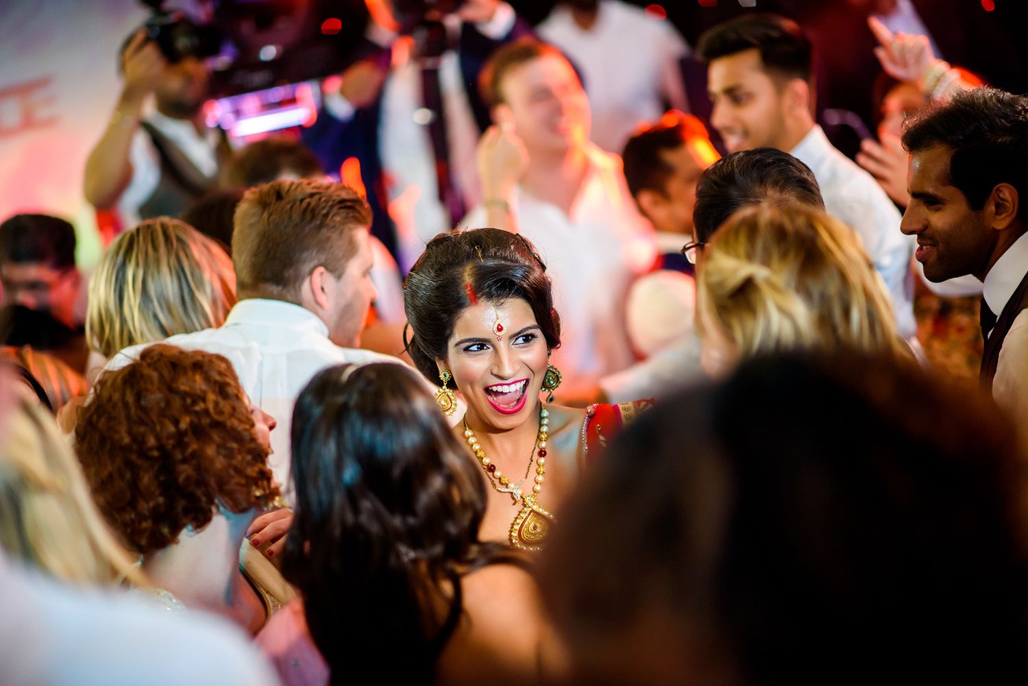 Crowded dance floor illuminated by warm pink lighting at an energetic wedding celebration.