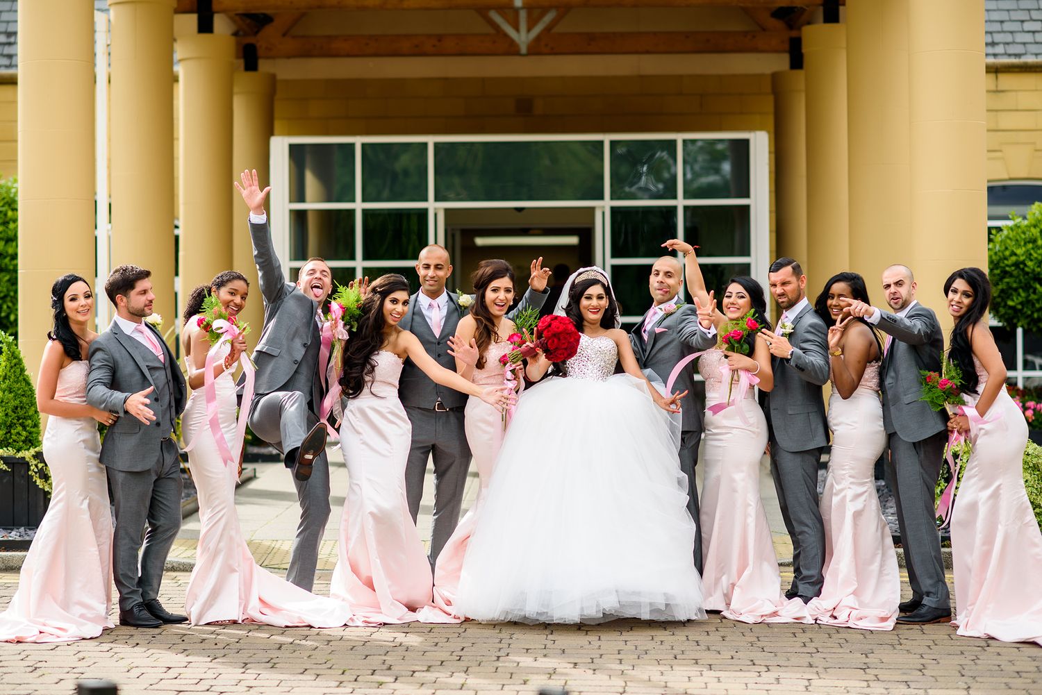 Wedding party stands in a line with bridesmaids in pink gowns outside a columned building.