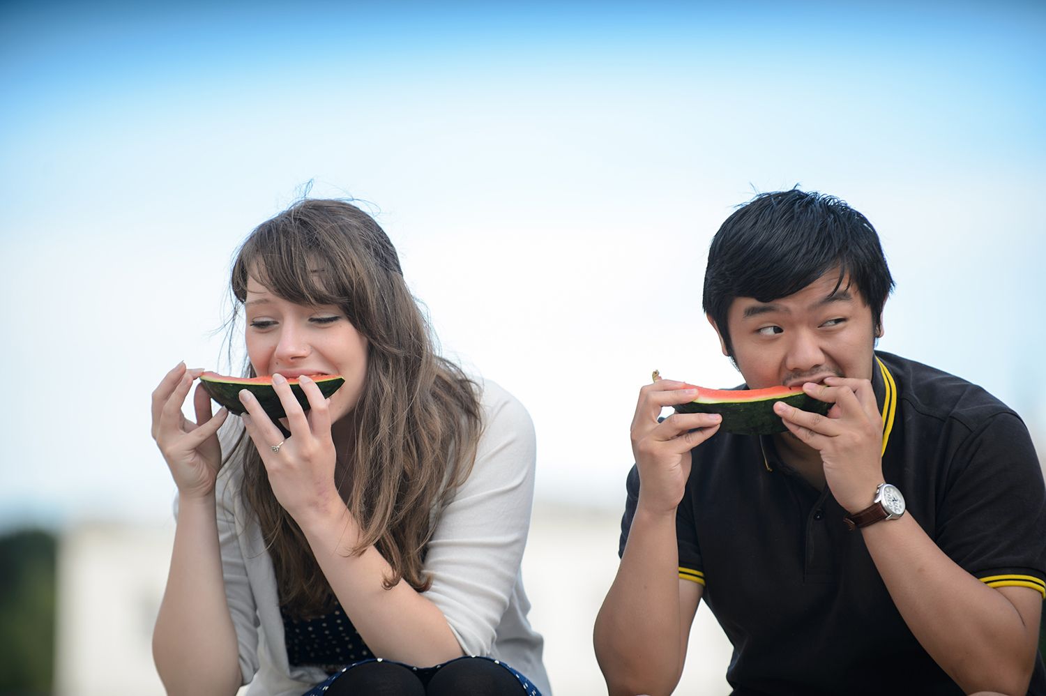 Two people laugh while eating snacks together outdoors near a historic building.