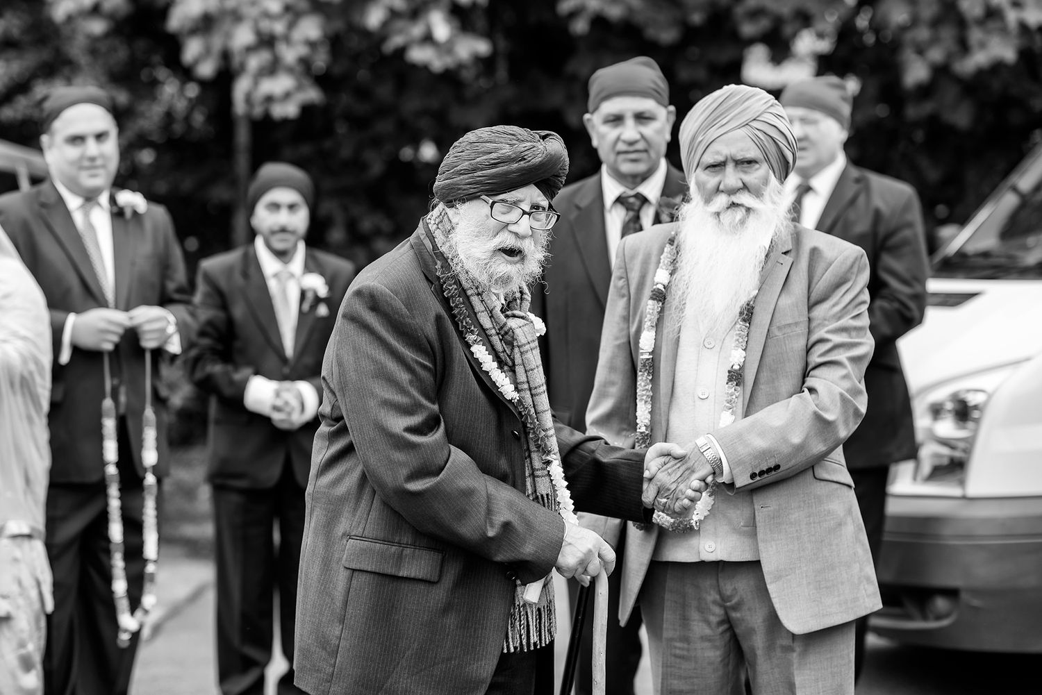Two elderly men in traditional attire shake hands at a formal outdoor gathering.