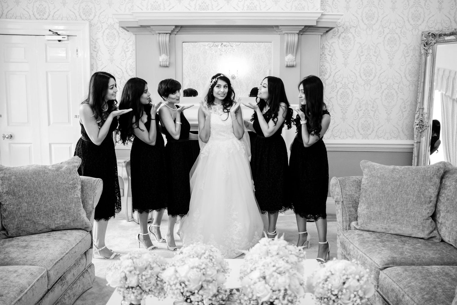 Wedding party lined up in formal attire against an ornate white wall backdrop.