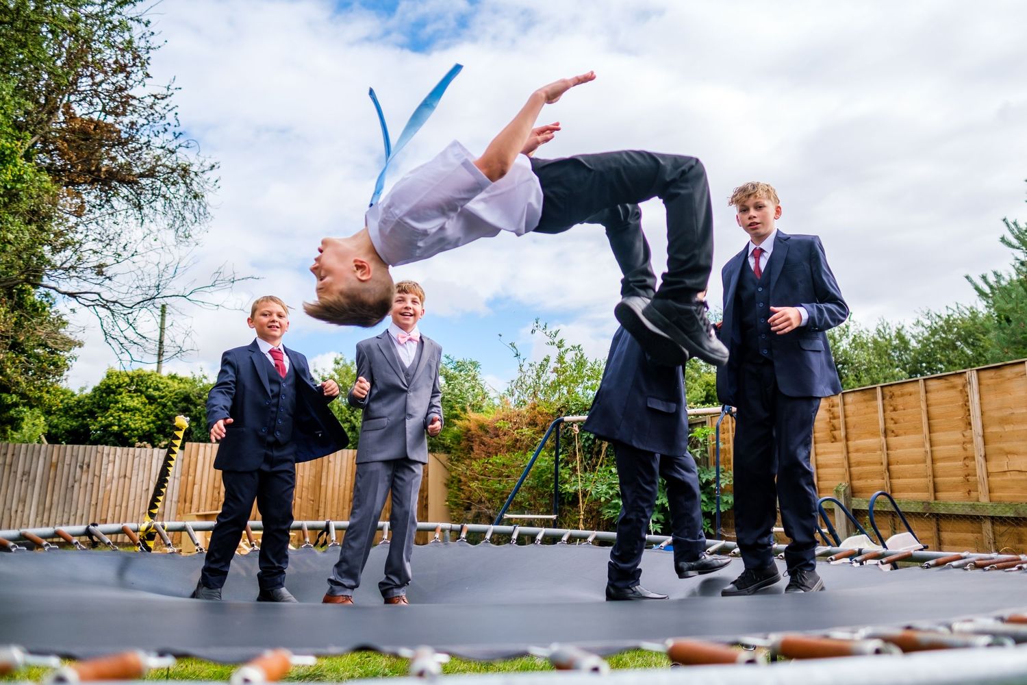 Person doing a backflip on a trampoline while others in suits watch in a backyard setting.