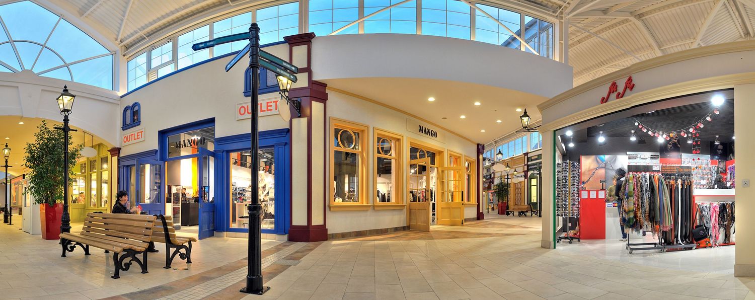 Modern shopping mall interior with curved glass ceiling, bright lighting and retail storefronts.