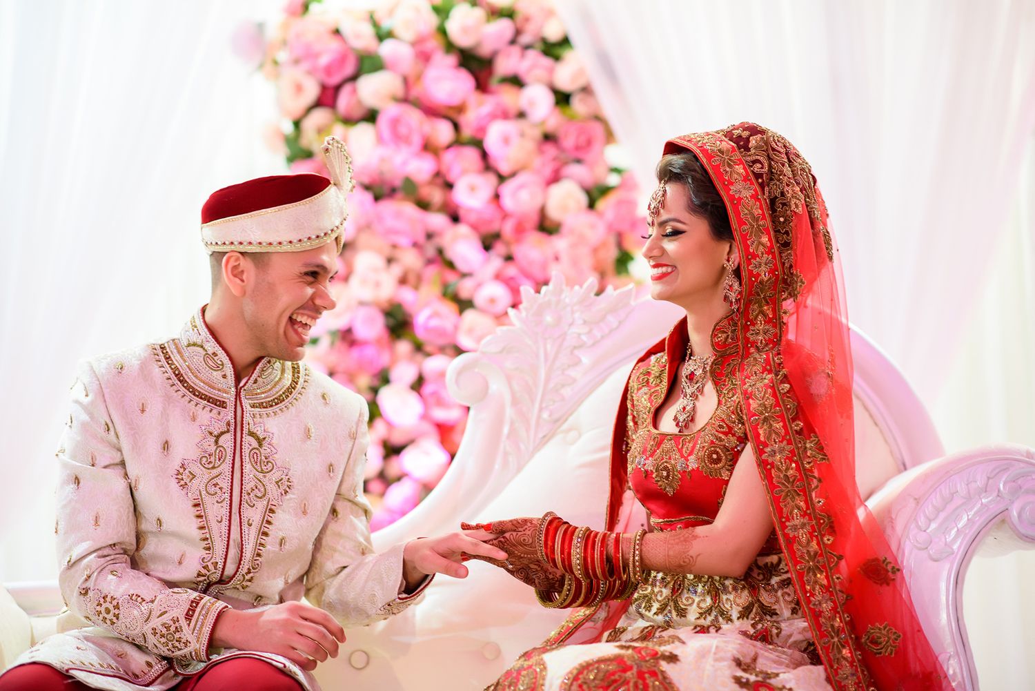 Wedding couple in traditional attire seated against pink floral backdrop during ceremony.