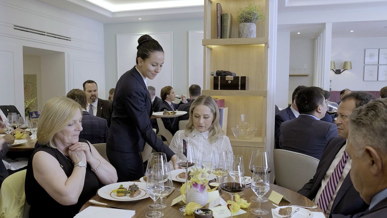 Elegant restaurant dining scene with a waitress serving guests at a formal table set with wine glasses and fine dining plates.