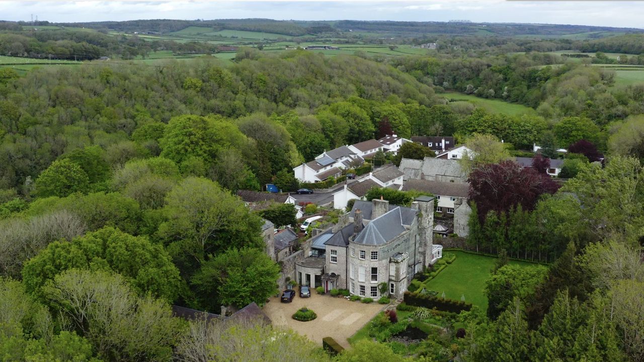 Aerial view of a grand stone country house surrounded by lush green trees and manicured gardens in a rural valley.