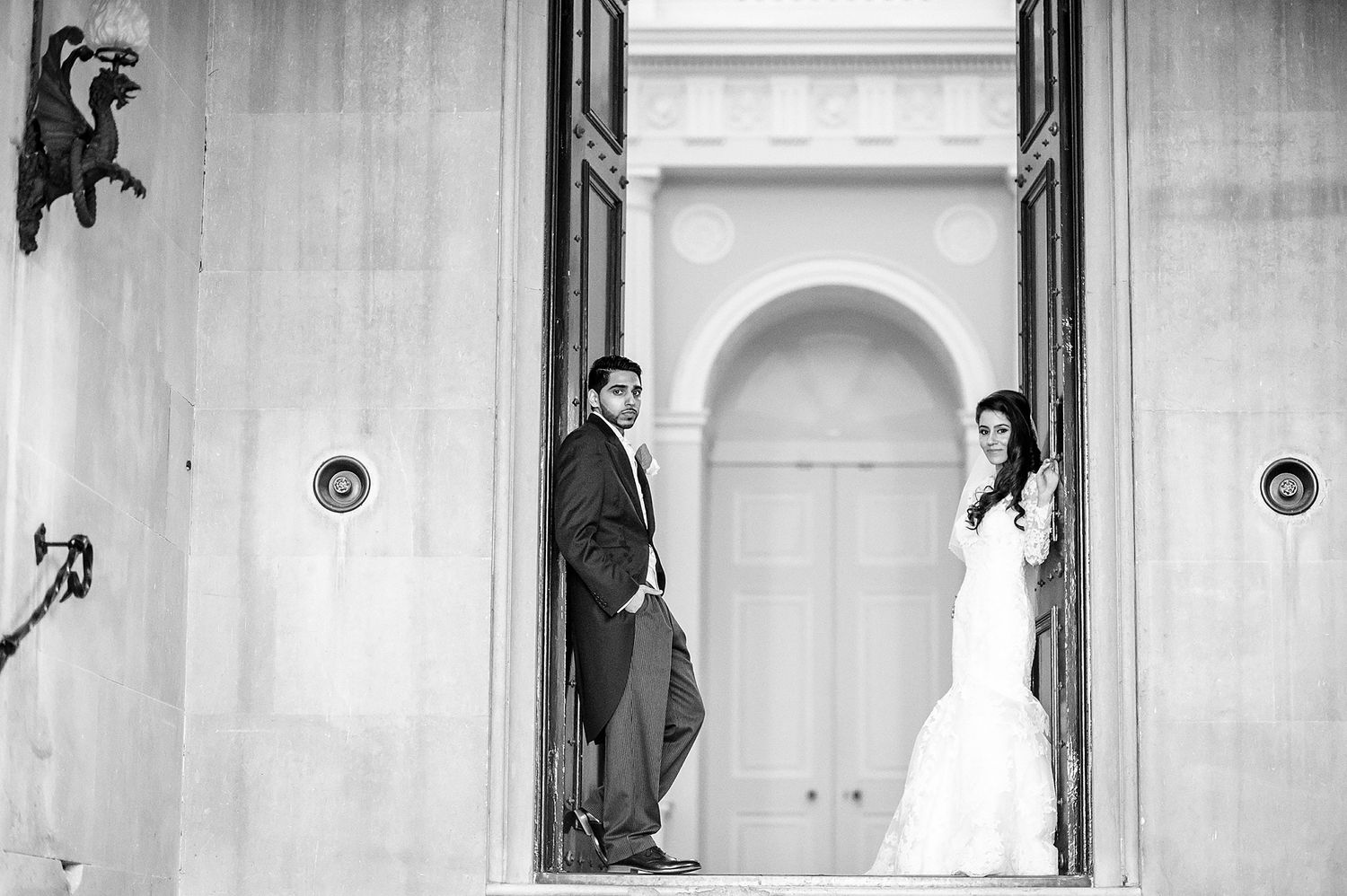 Black and white photo of couple posing in arched doorway wearing formal wedding attire.
