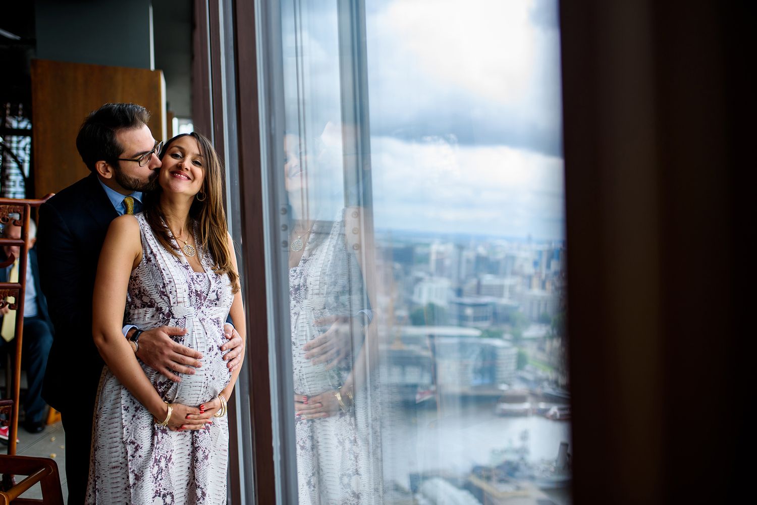A couple stands intimately by floor-to-ceiling windows overlooking a city vista.