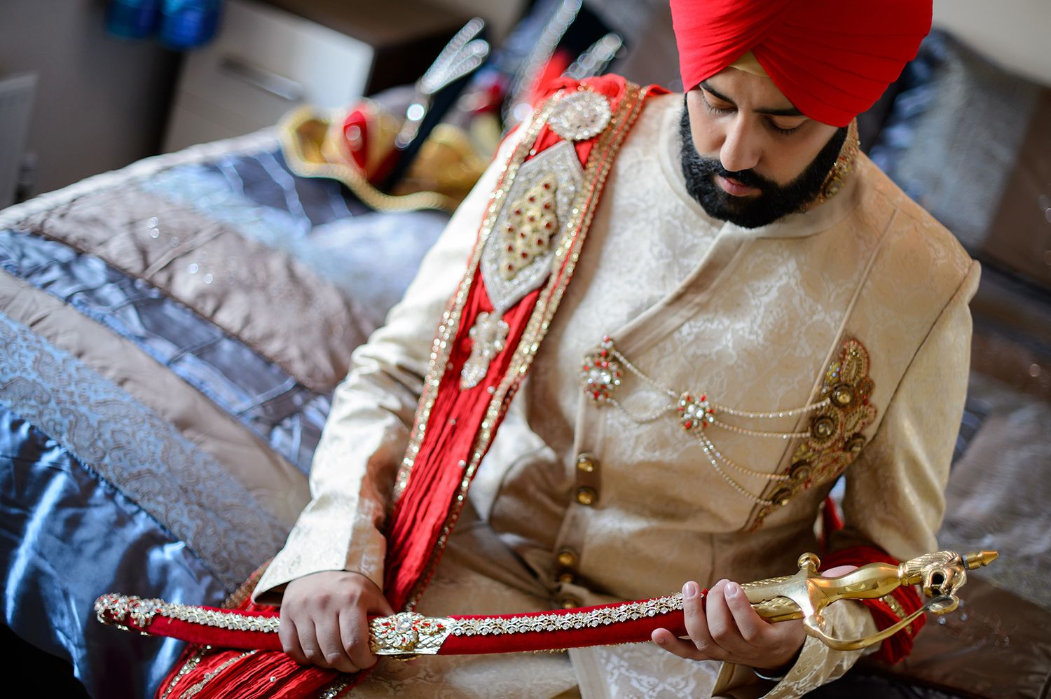 Traditional Sikh groom in red turban and cream sherwani holding ceremonial sword.