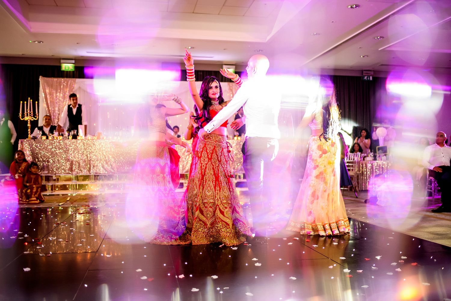 Wedding guests dancing and celebrating at a vibrant reception with purple and pink lighting effects.