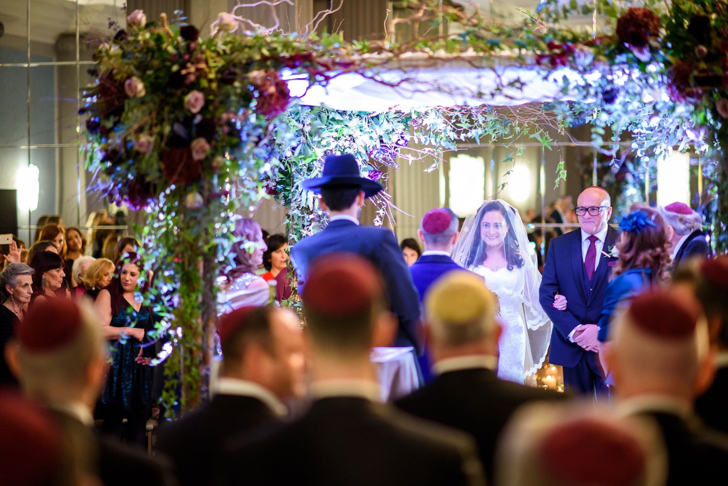 Wedding ceremony under a glowing blue and purple floral arch with guests gathered in formal attire.