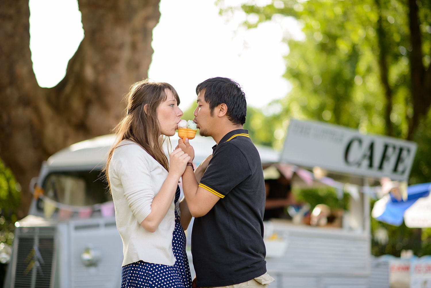 Two people share ice cream cones near a cafe cart on a sunny day.