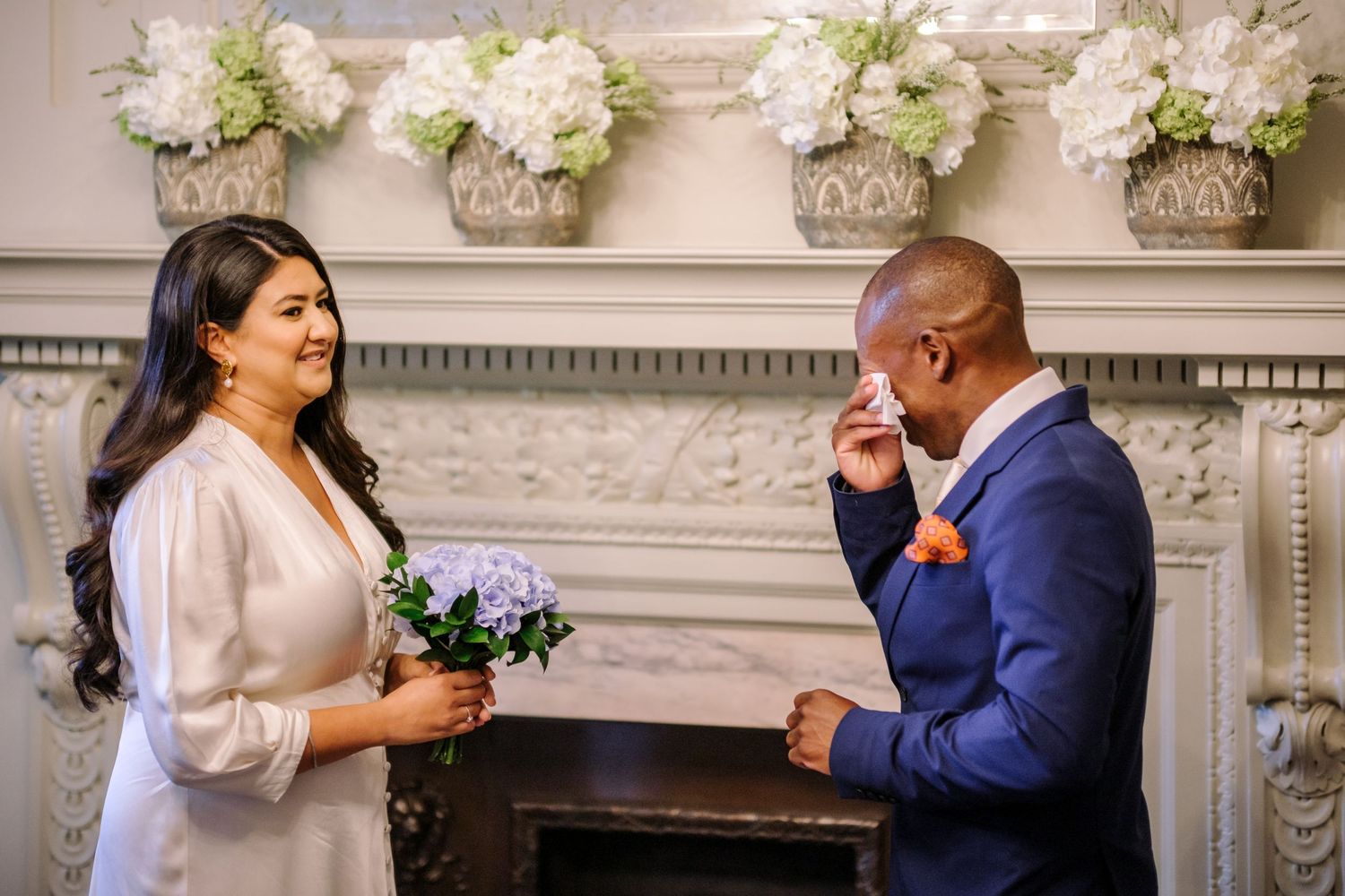 An emotional wedding moment captured in front of an elegant fireplace decorated with white floral arrangements.