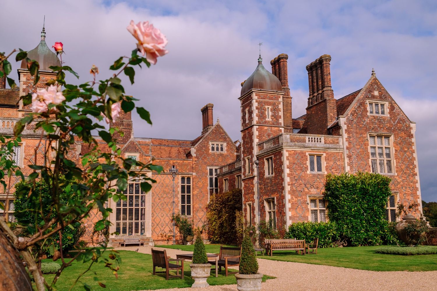 Historic English manor house with red brick facade and ornate chimneys surrounded by manicured gardens and a pink rose.