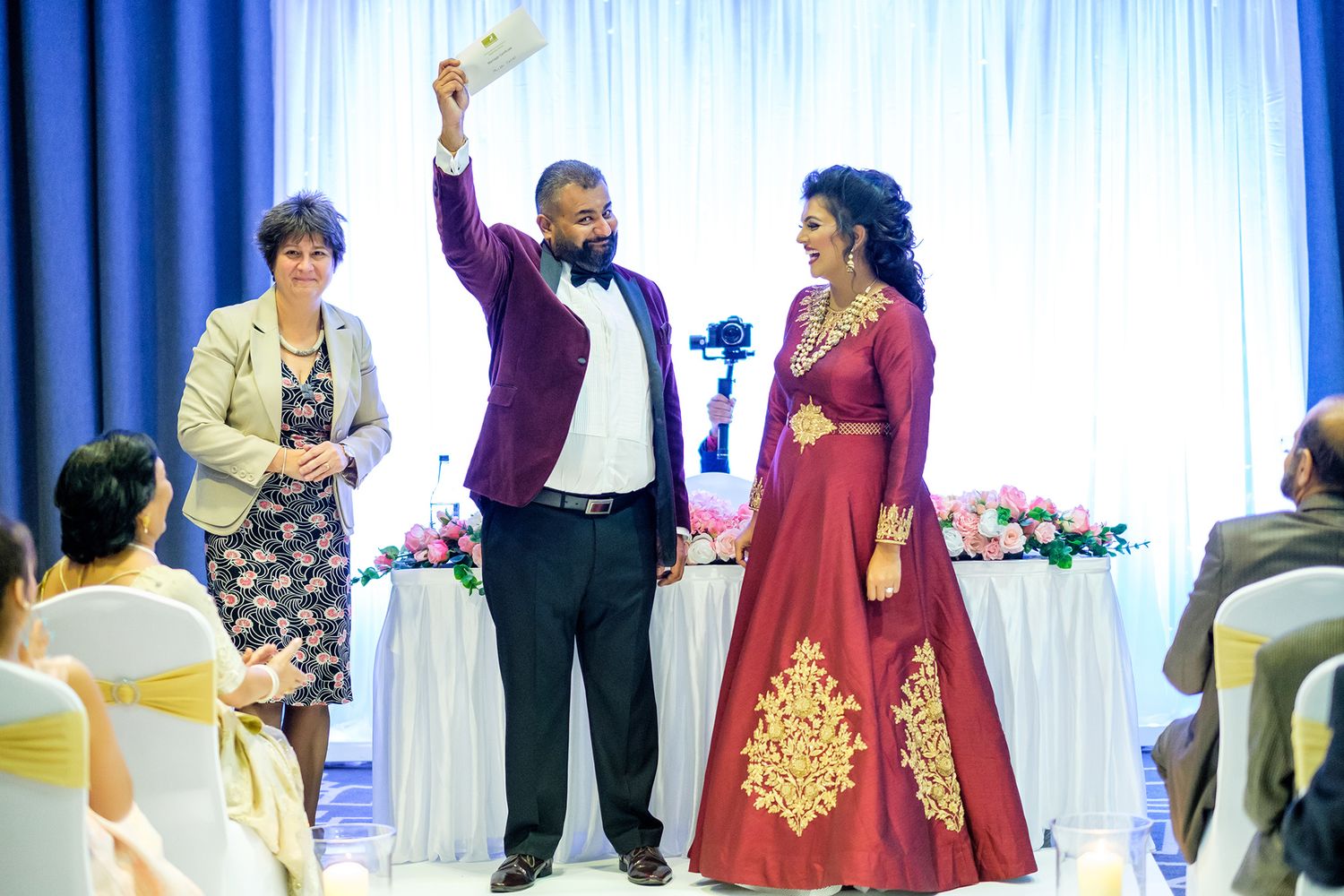 Wedding couple making their entrance with raised arms in front of decorated white backdrop.