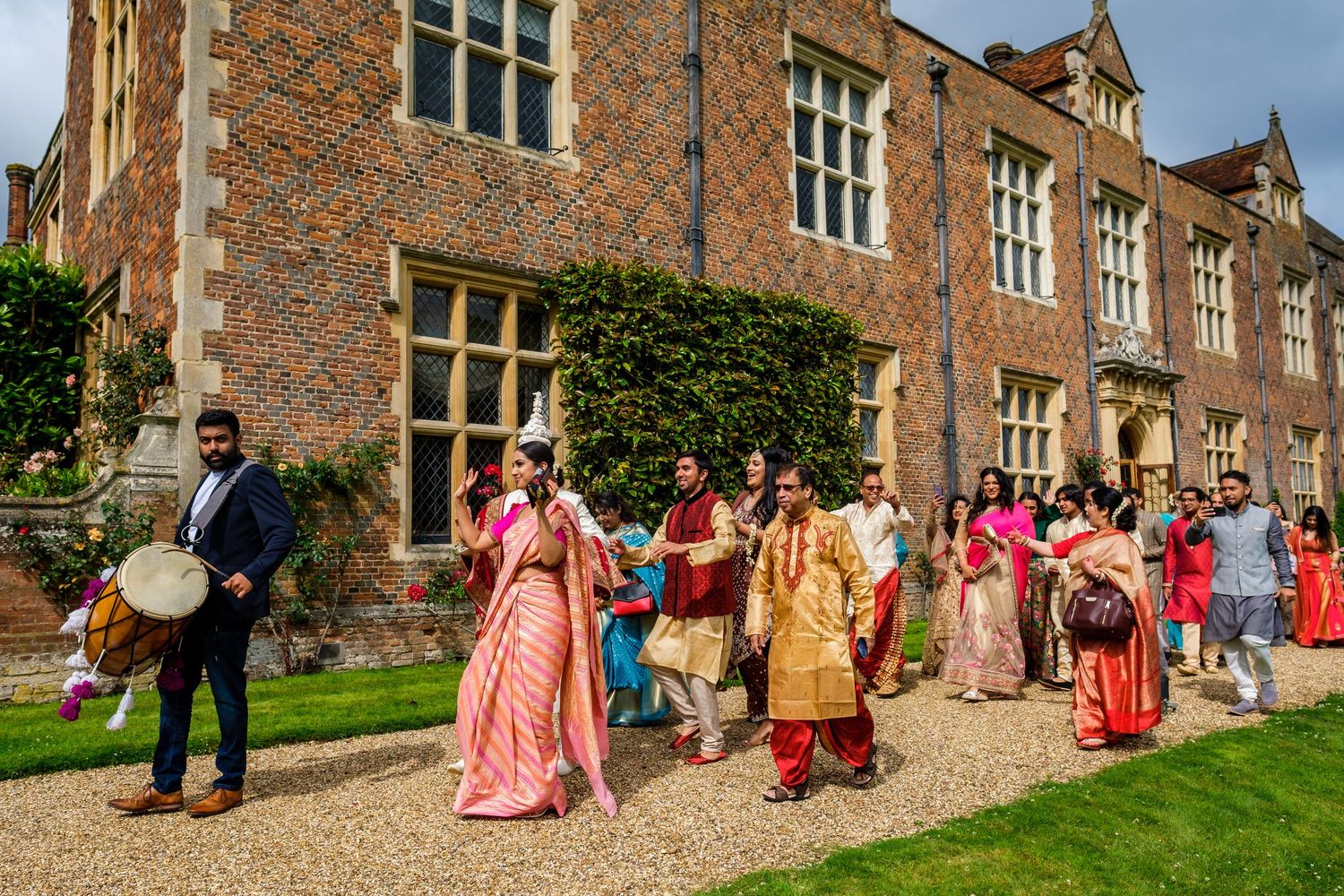 A festive procession of guests in colorful traditional Indian attire walk along a historic brick manor house.