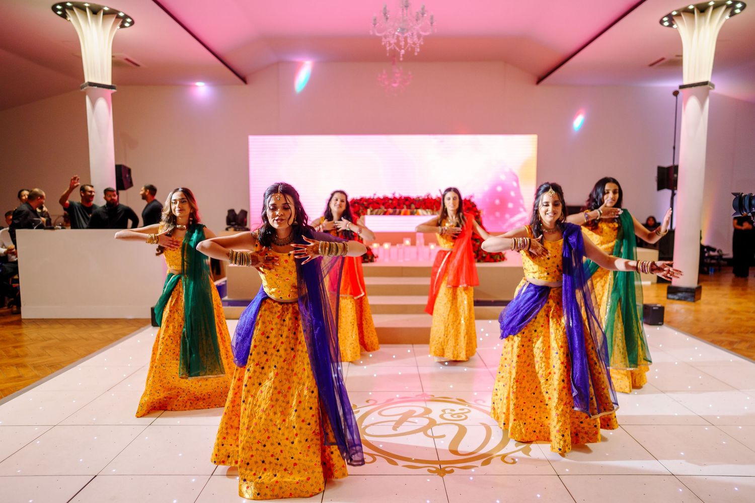 Traditional Indian dance performance with dancers in vibrant yellow and blue lehengas on a white stage with pink lighting.