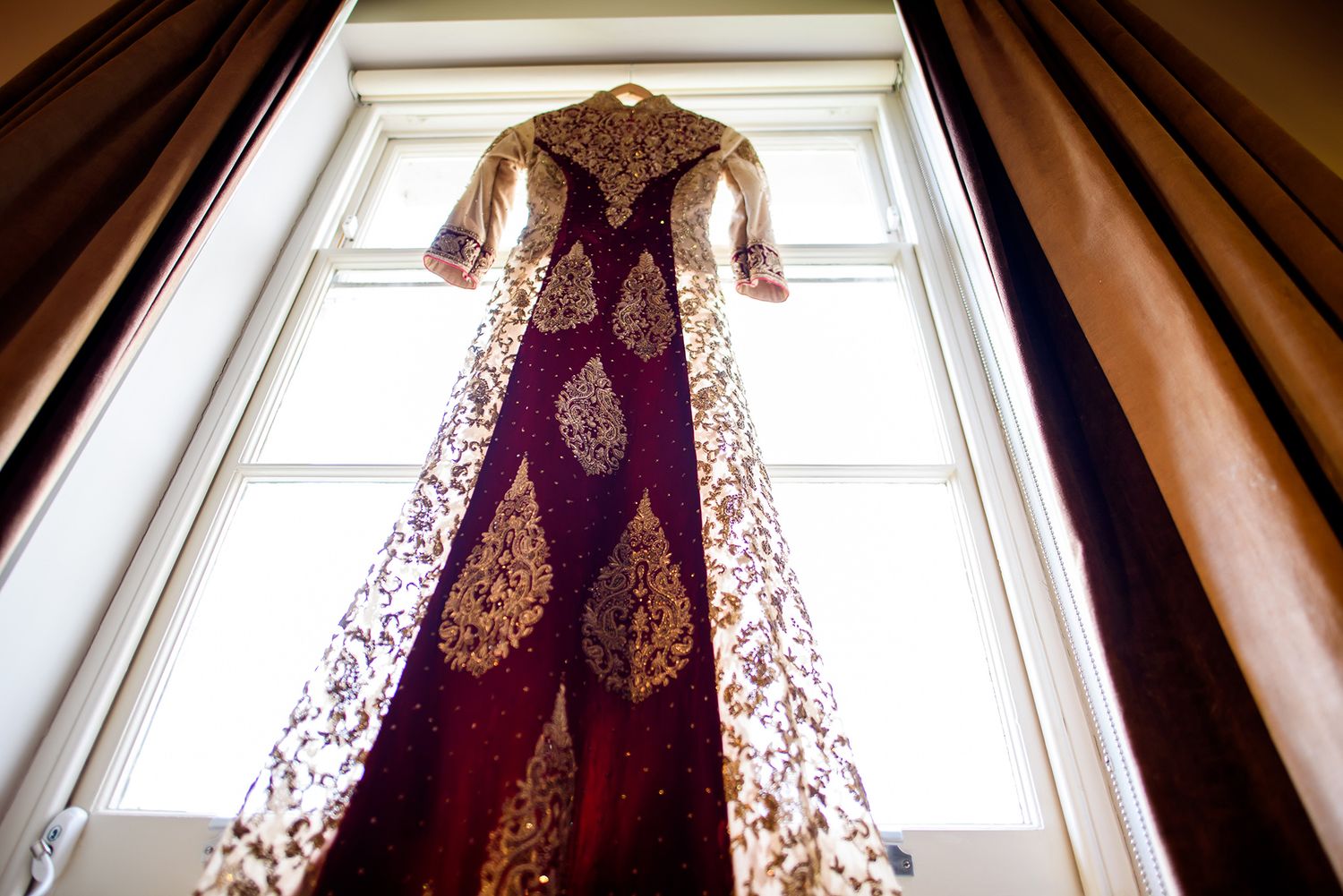 Ornate burgundy and gold wedding dress displayed in front of sunlit window.