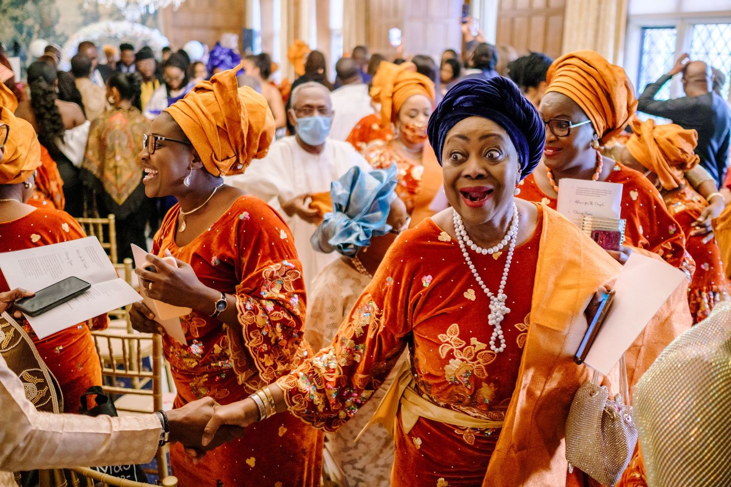 Wedding guests in vibrant orange and red traditional attire celebrate joyfully during reception.