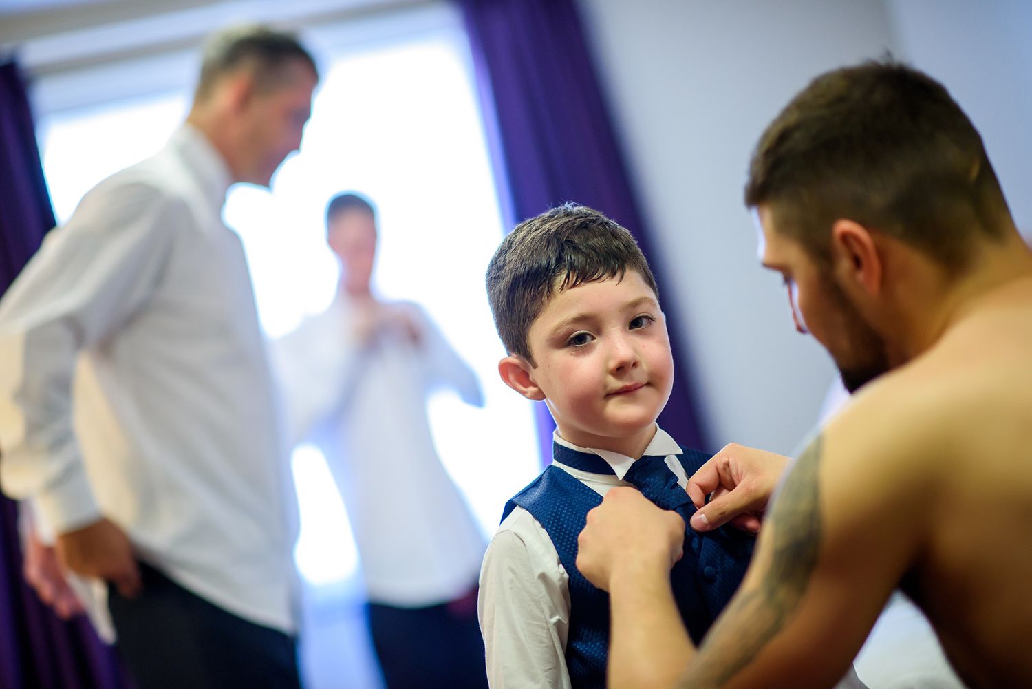 A young child having a tie adjusted while others prepare in the background.