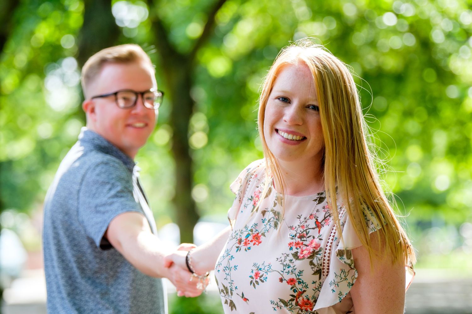 A couple poses outdoors in a park with vibrant green foliage in the background.