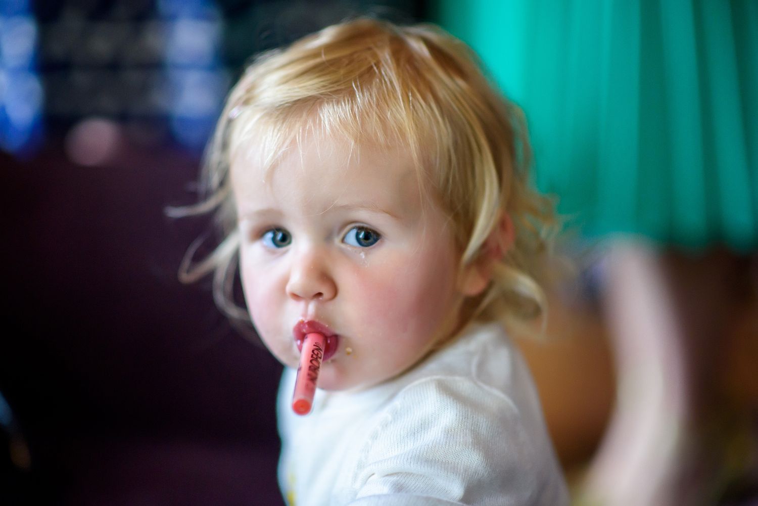 Toddler with blonde hair enjoys a lollipop against a turquoise background.