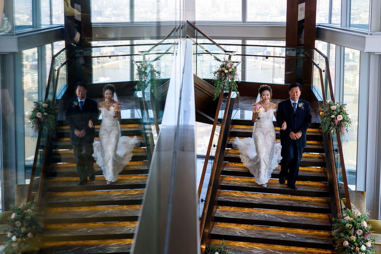 Couple poses on elegant spiral staircase decorated with greenery and white floral arrangements during wedding photoshoot.