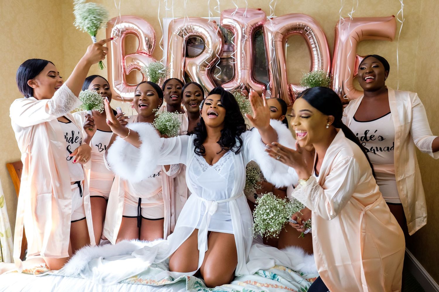 Bridal party in white robes celebrates on a bed with balloon letters spelling BRIDE.