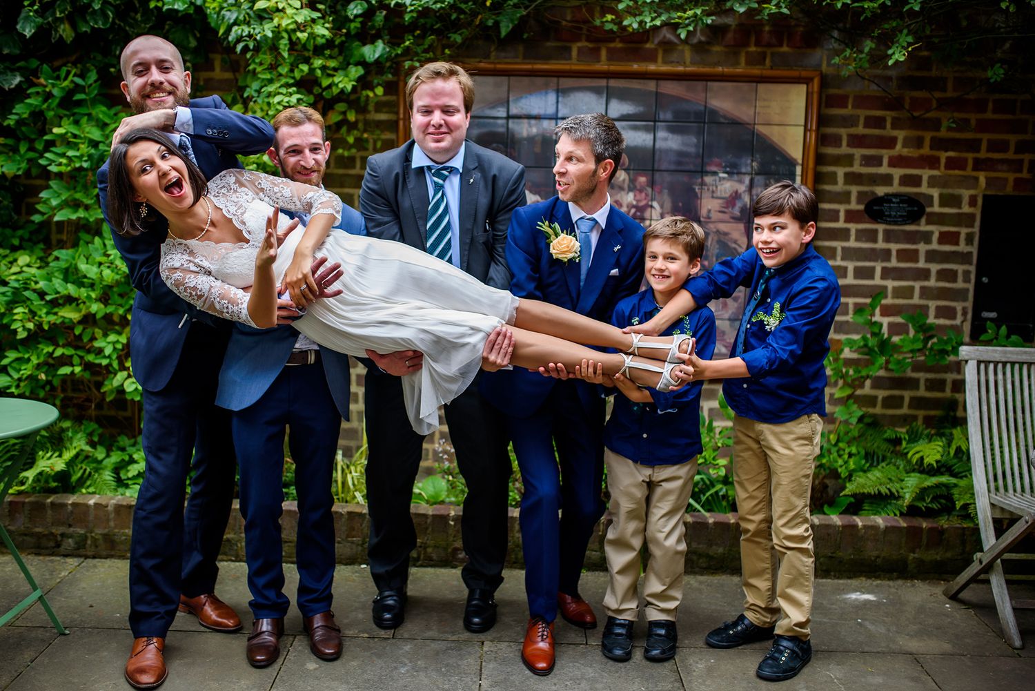 Groomsmen in navy suits playfully lift and carry someone in a fun wedding party photo outdoors.