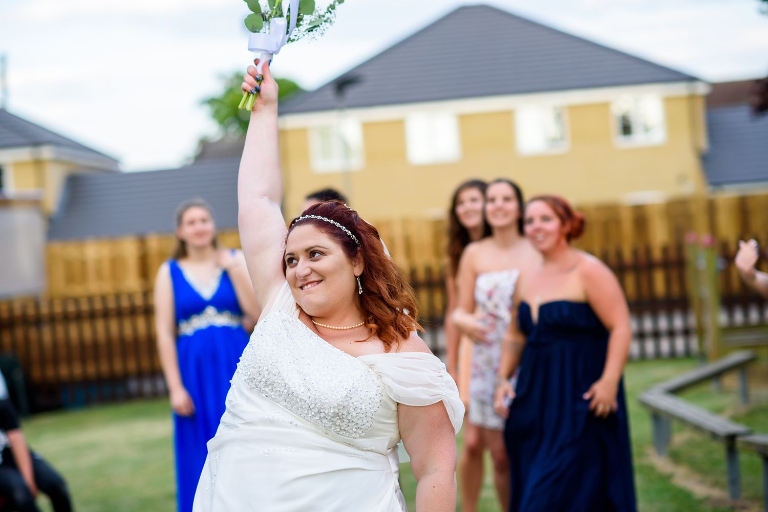 Wedding party celebrates on lawn with yellow house and wooden fence in background.