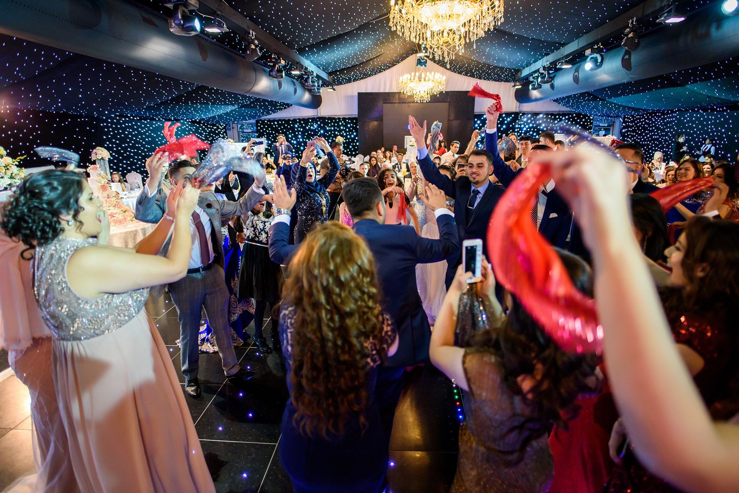 Wedding reception party crowd dancing under a crystal chandelier and twinkling lights.