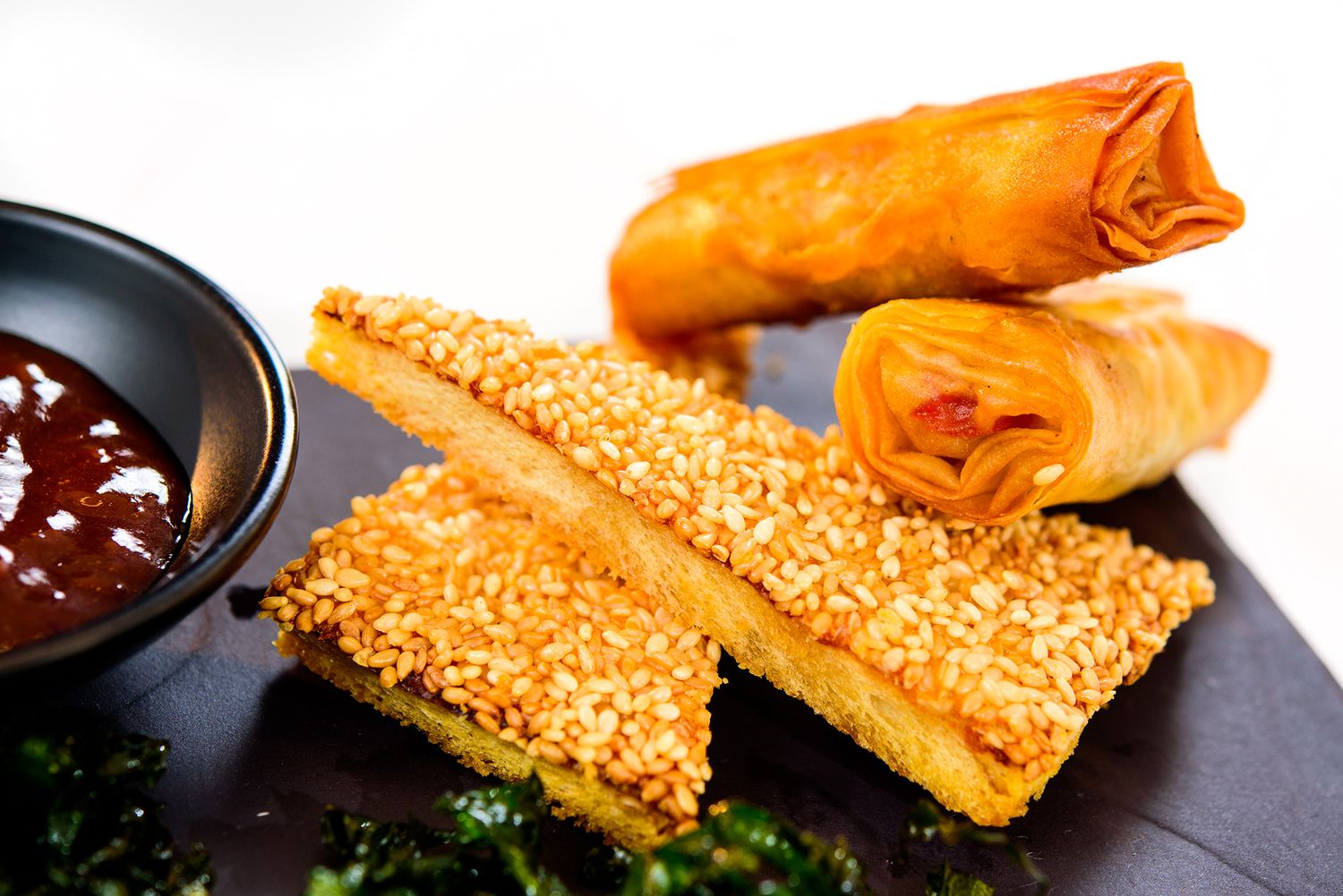 Assorted Asian appetizers including spring rolls and sesame-crusted items served with dipping sauces on a dark platter.