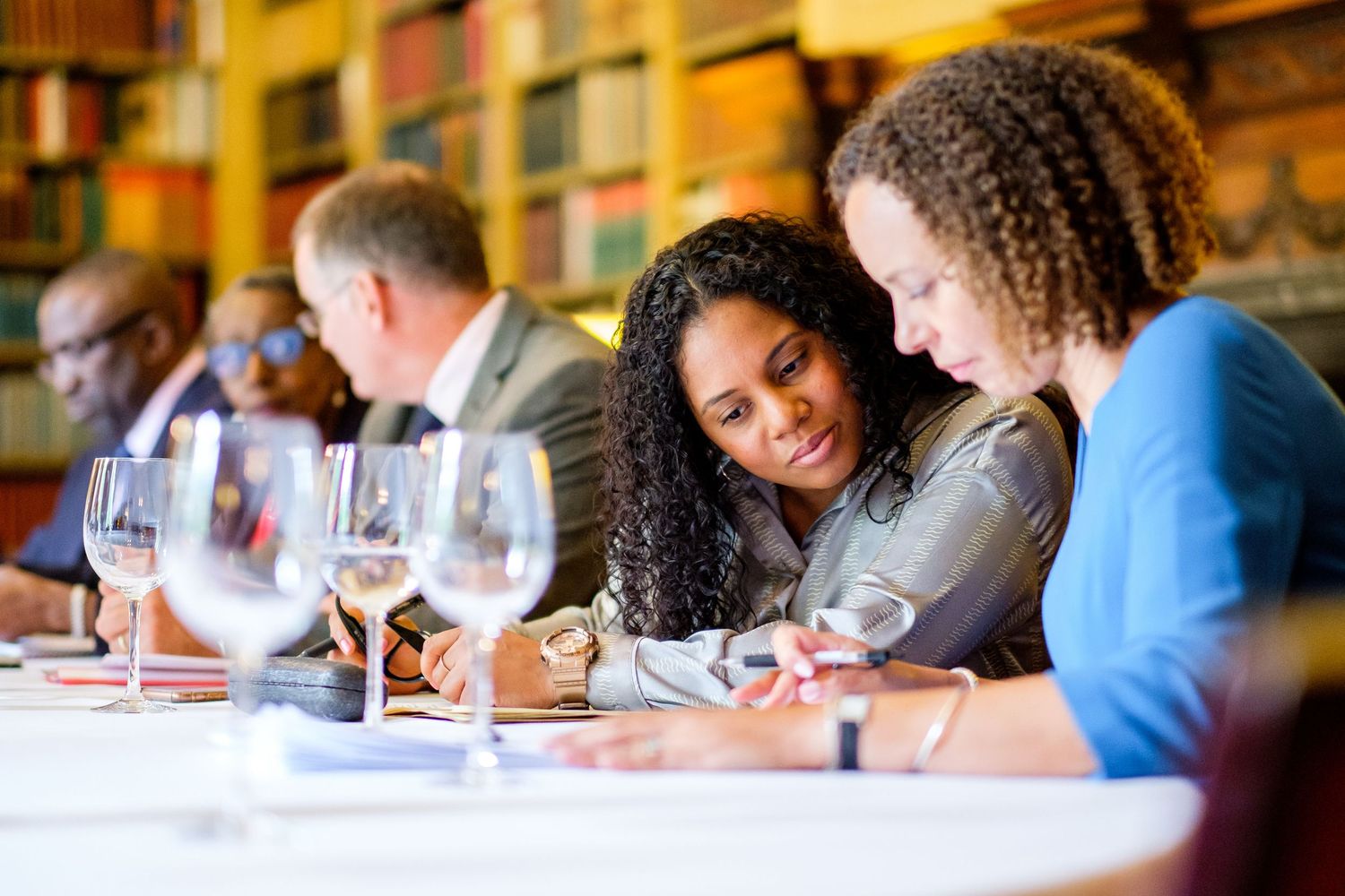 Group tasting wine at wooden table in library setting.