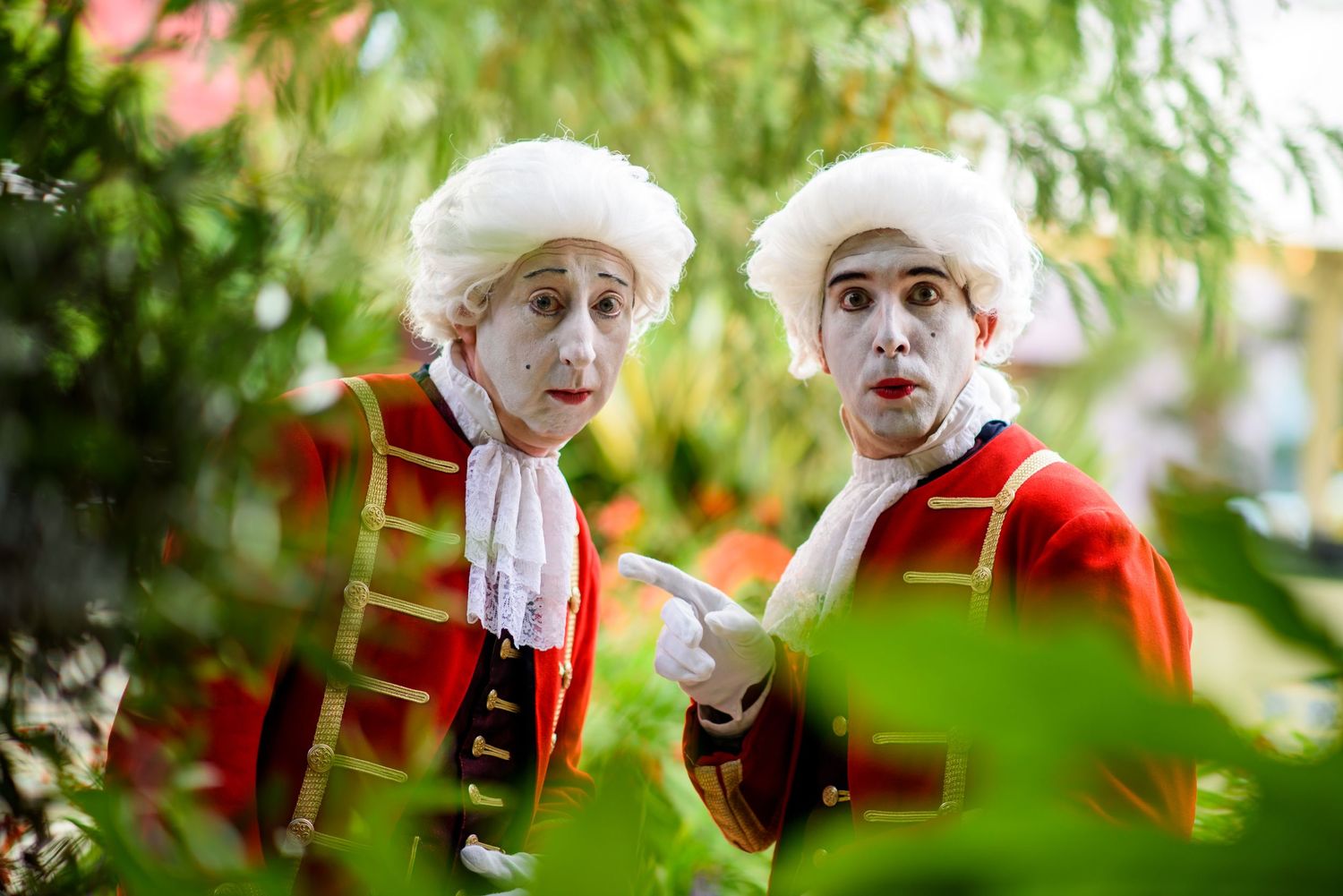 A person in a red dress and white wig performs expressively against a green natural outdoor background.
