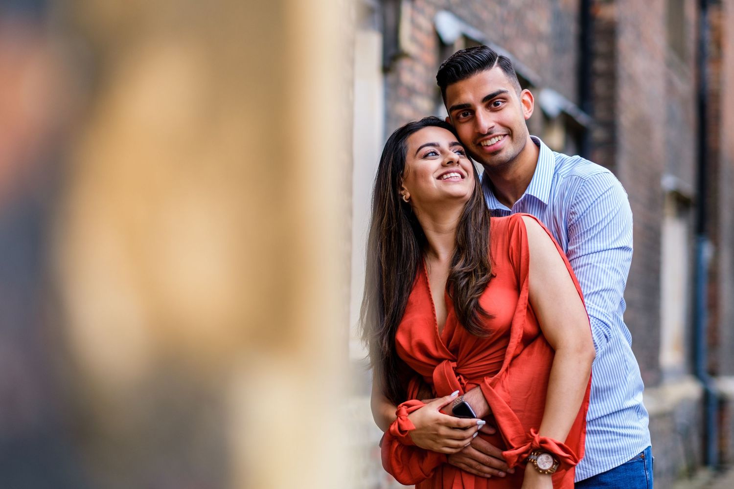 A couple in casual attire share romantic moments while posing against a rustic brick wall background.